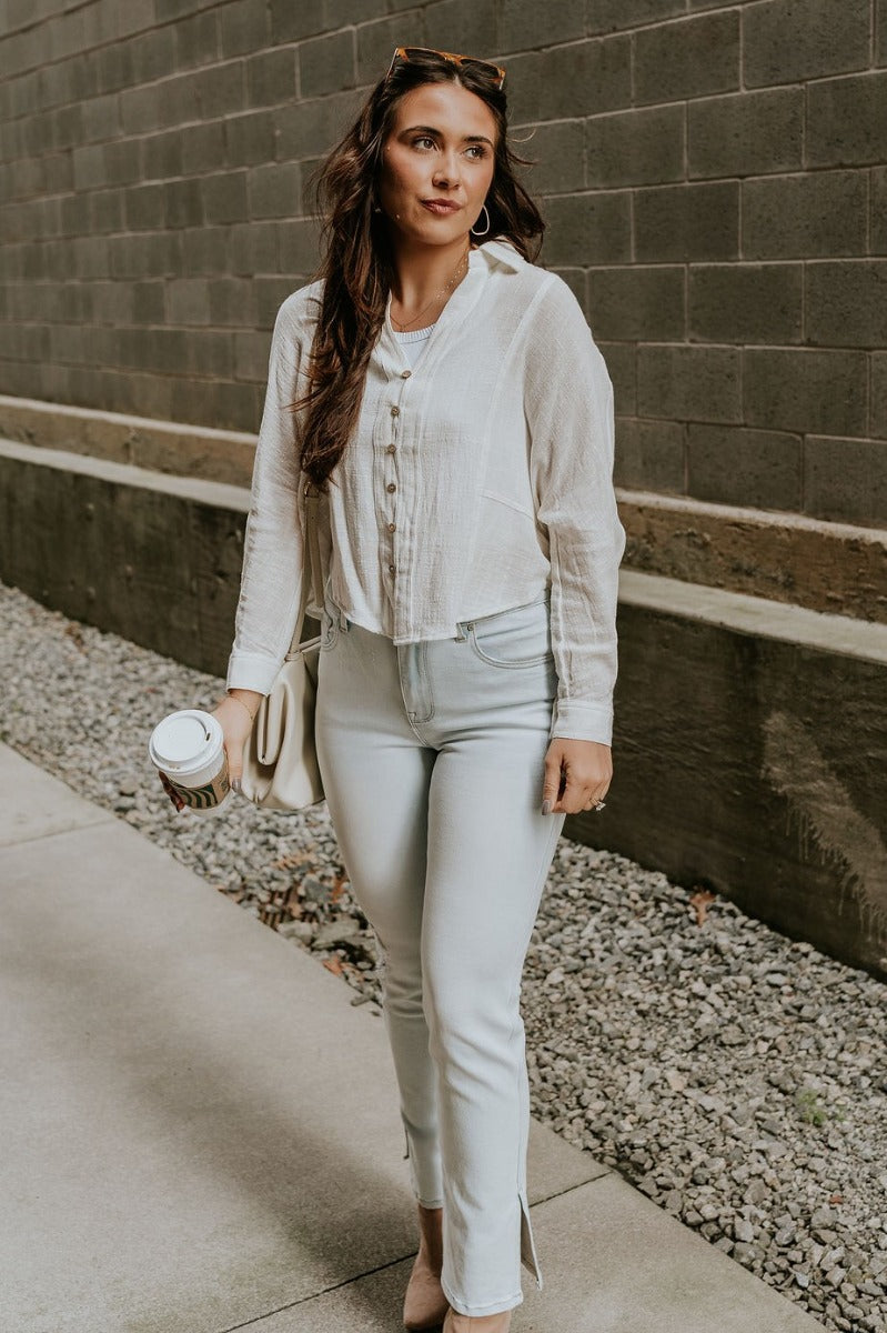 Full body front view of model wearing the Elisabeth White Gauze Long Sleeve Button Up Top that has off white textured fabric, wooden buttons, a collar, stitched hem details and long sleeves.