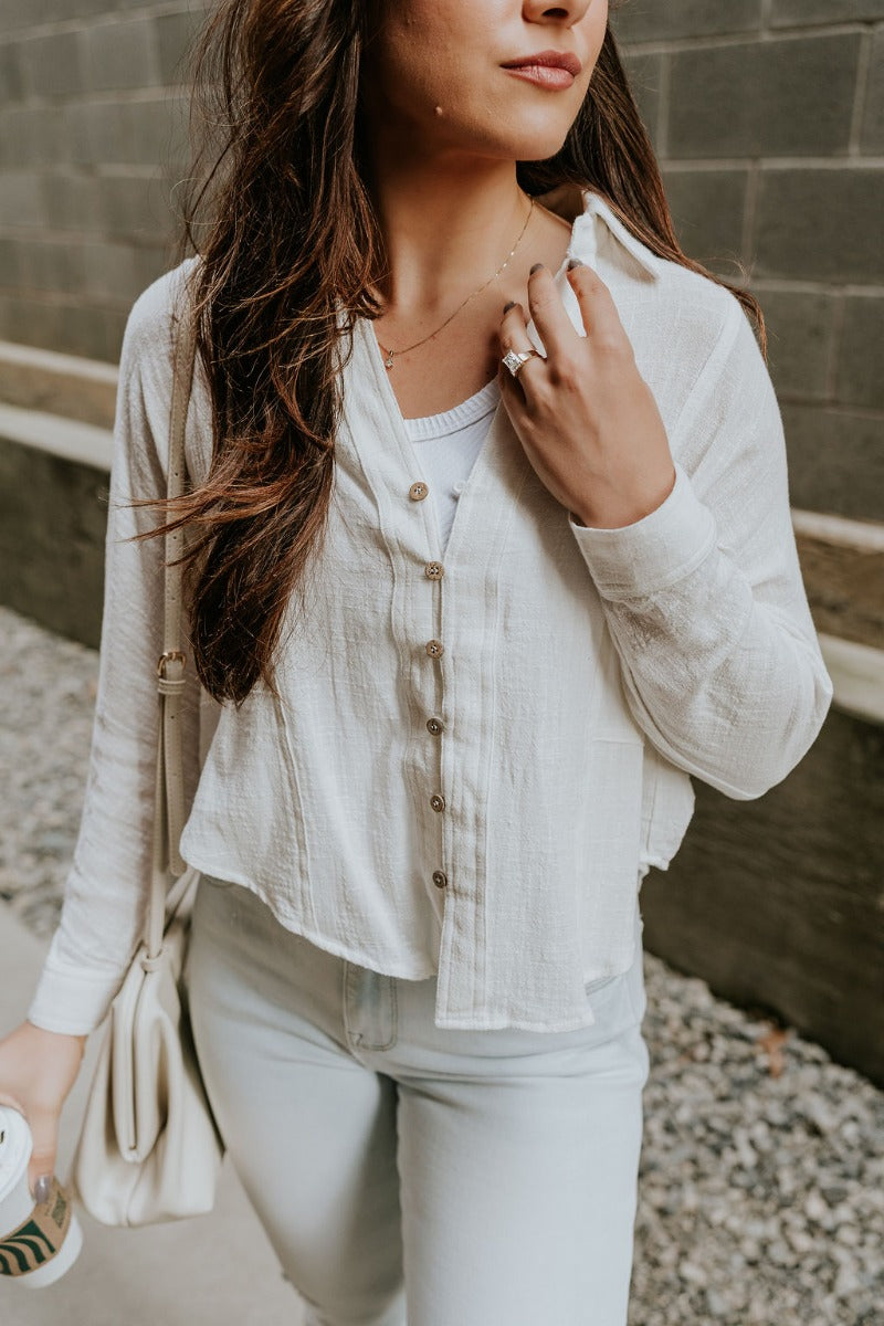 Close front view of model wearing the Elisabeth White Gauze Long Sleeve Button Up Top that has off white textured fabric, wooden buttons, a collar, stitched hem details and long sleeves.