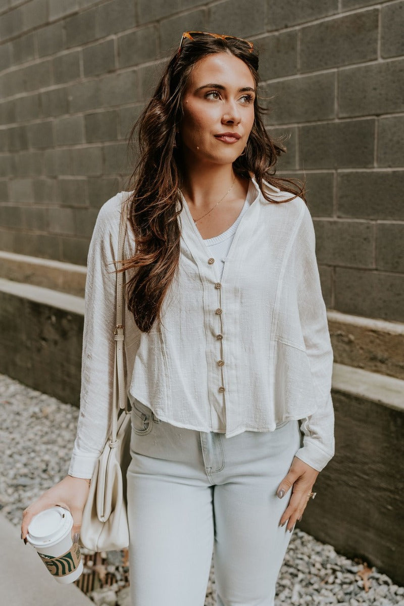 front view of model wearing the Elisabeth White Gauze Long Sleeve Button Up Top that has off white textured fabric, wooden buttons, a collar, stitched hem details and long sleeves.