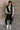 Full body front view of model wearing the Camryn Black Longline Puffer Vest features black puffer fabric, midi length, pockets, a hidden zipper with snap closures, a high neck, and a hood with drawstring ties.