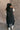 Full body back view of model wearing the Camryn Black Longline Puffer Vest features black puffer fabric, midi length, pockets, a hidden zipper with snap closures, a high neck, and a hood with drawstring ties.