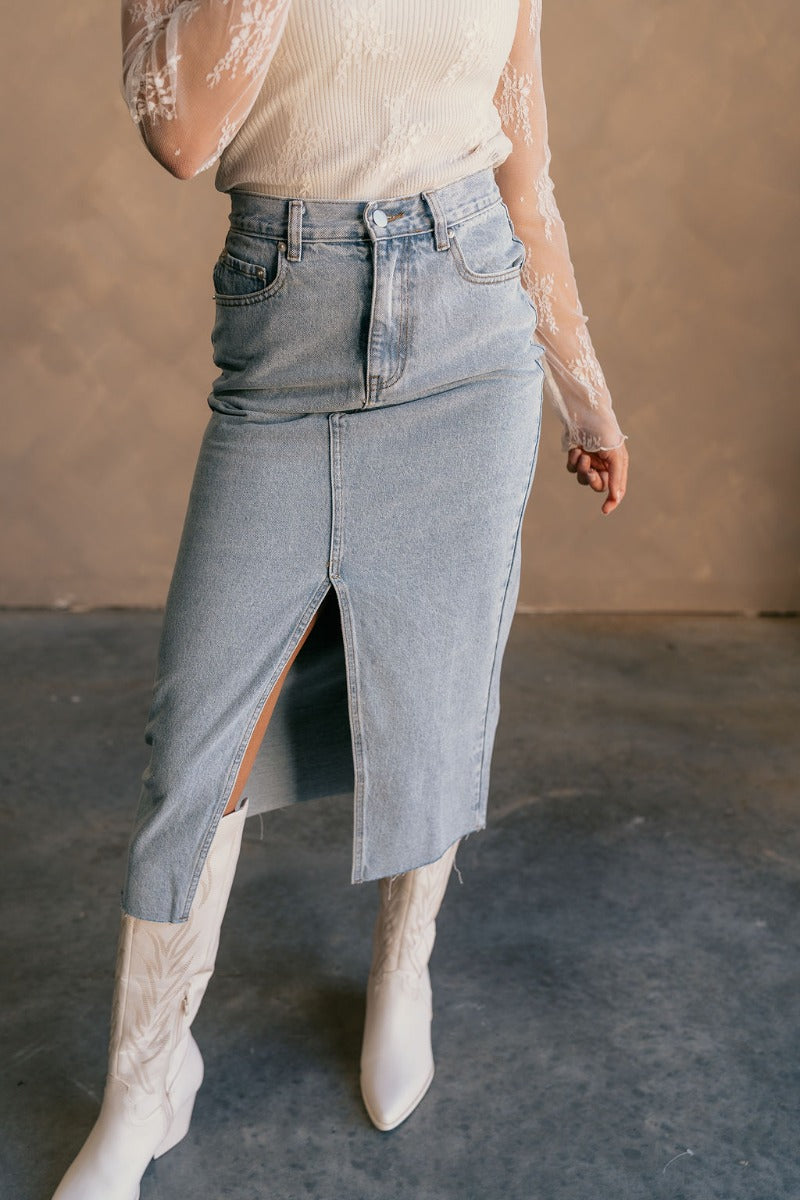 front view of model wearing the Liana Denim Front Slit Midi Skirt that has light wash denim fabric, brown stitching, midi length, a raw hem, and a front slit.