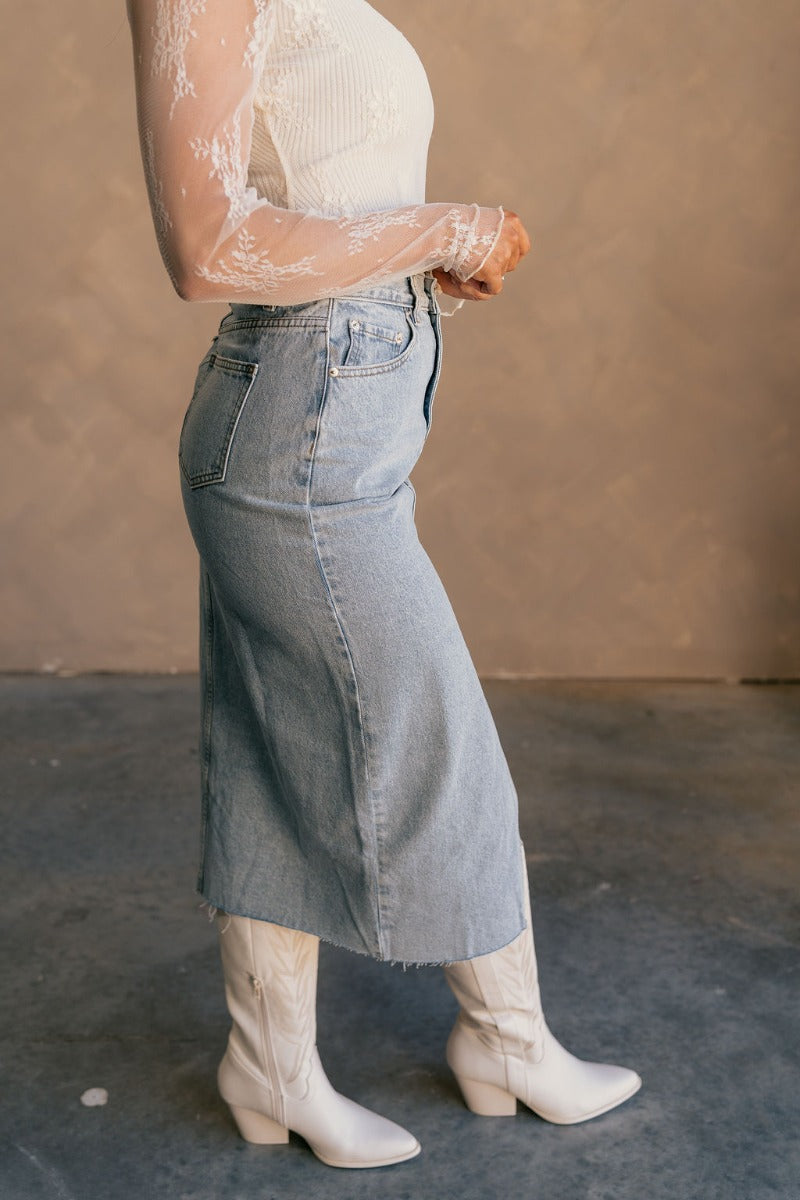Side view of model wearing the Liana Denim Front Slit Midi Skirt that has light wash denim fabric, brown stitching, midi length, a raw hem, and a front slit.