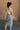 Full body back view of model wearing the Liana Denim Front Slit Midi Skirt that has light wash denim fabric, brown stitching, midi length, a raw hem, and a front slit.