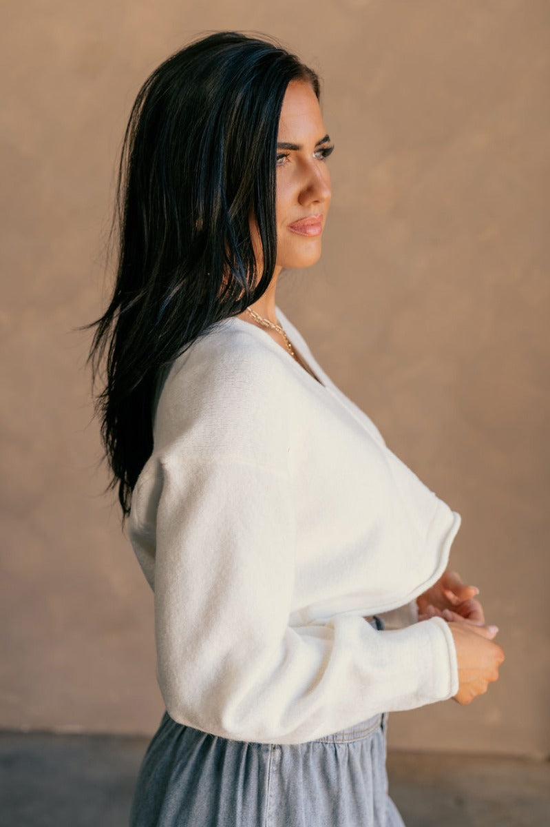 Side view of model wearing the Angela Ivory Knit long Sleeve Sweater which features off white knit fabric, a cropped waist, a v-neckline, and long sleeves.
