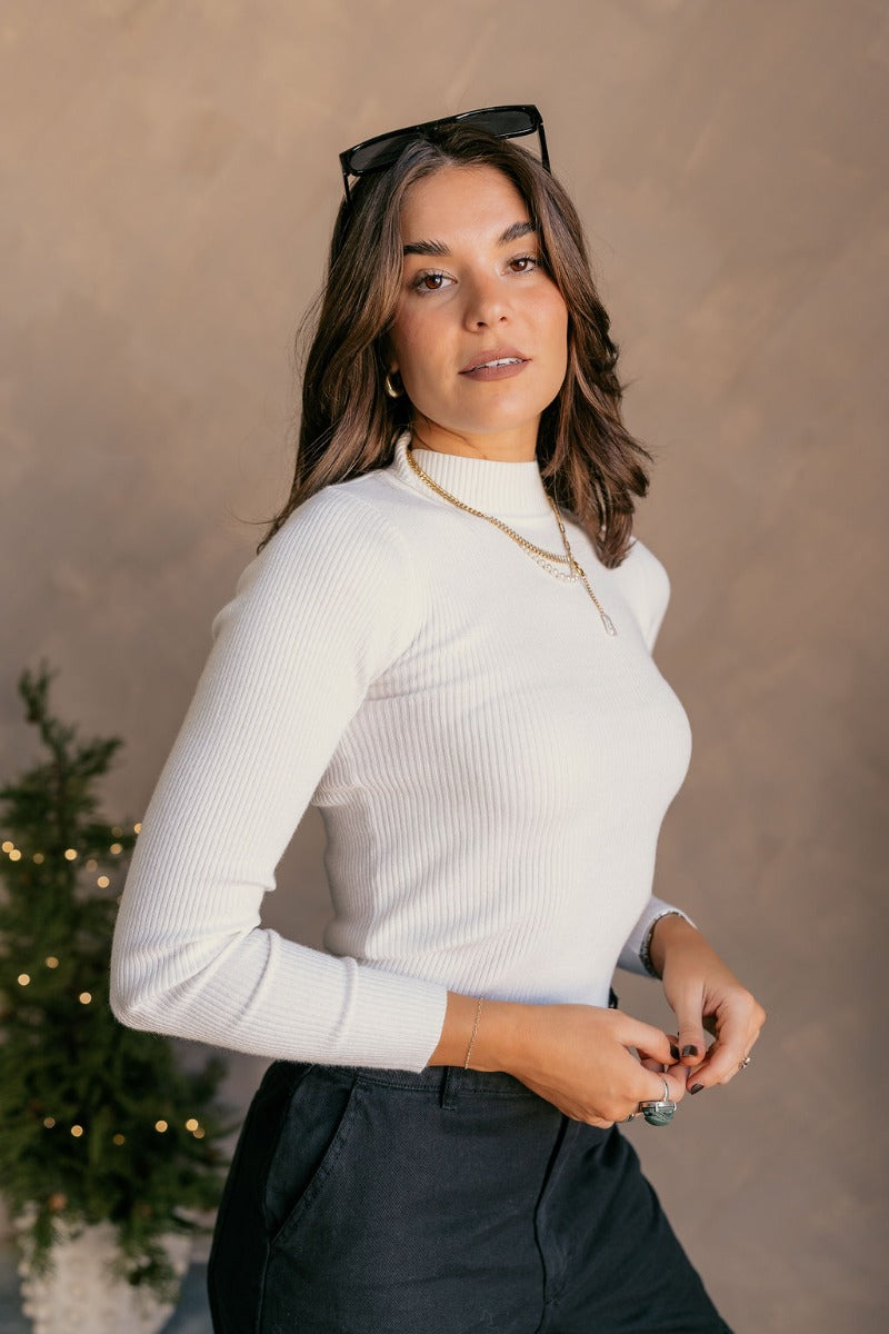 Side view of model wearing the Mila Ivory Ribbed Long Sleeve Top which features ivory ribbed knit fabric, a high neckline, and long sleeves.