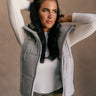 Front view of model wearing the Leah Heather Grey Hoodie Vest which features heather grey knit puffer fabric, a drawstring hem, a front zipper, two front zip up pockets, a high neckline, and a hood with drawstring ties.