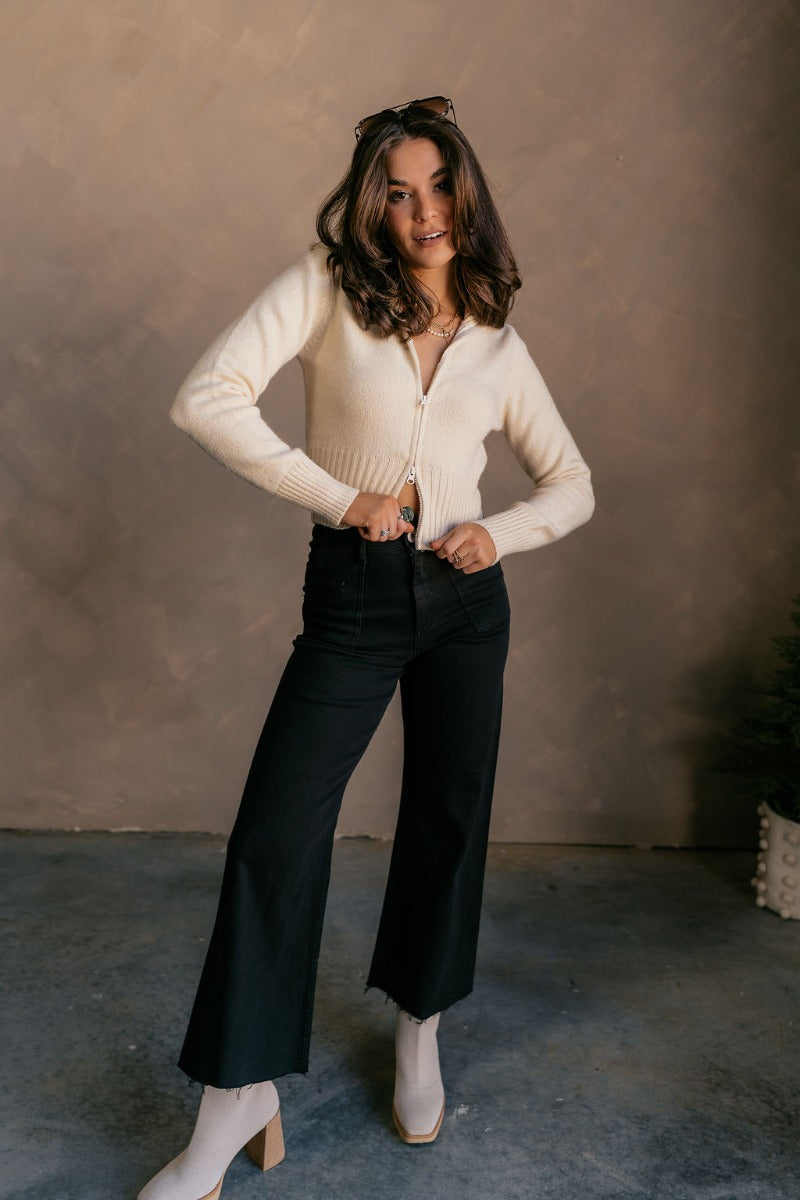Full body view of model wearing the Everly Beige Zip-Up Long Sleeve Sweater which features cream knit fabric, ribbed trim, a cropped waist, a monochrome front zipper, an adjustable neckline, and long sleeves with ribbed cuffs.