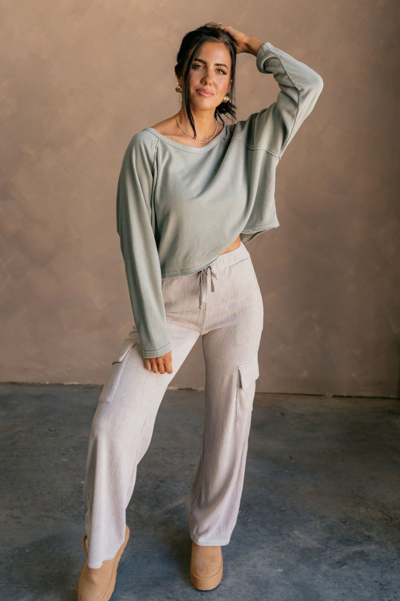 Full body front view of model wearing the Charlotte Sage Green Raglan Sweatshirt that has dusty light sage knit fabric, raw hem details, a round neckline, and long wide sleeves.