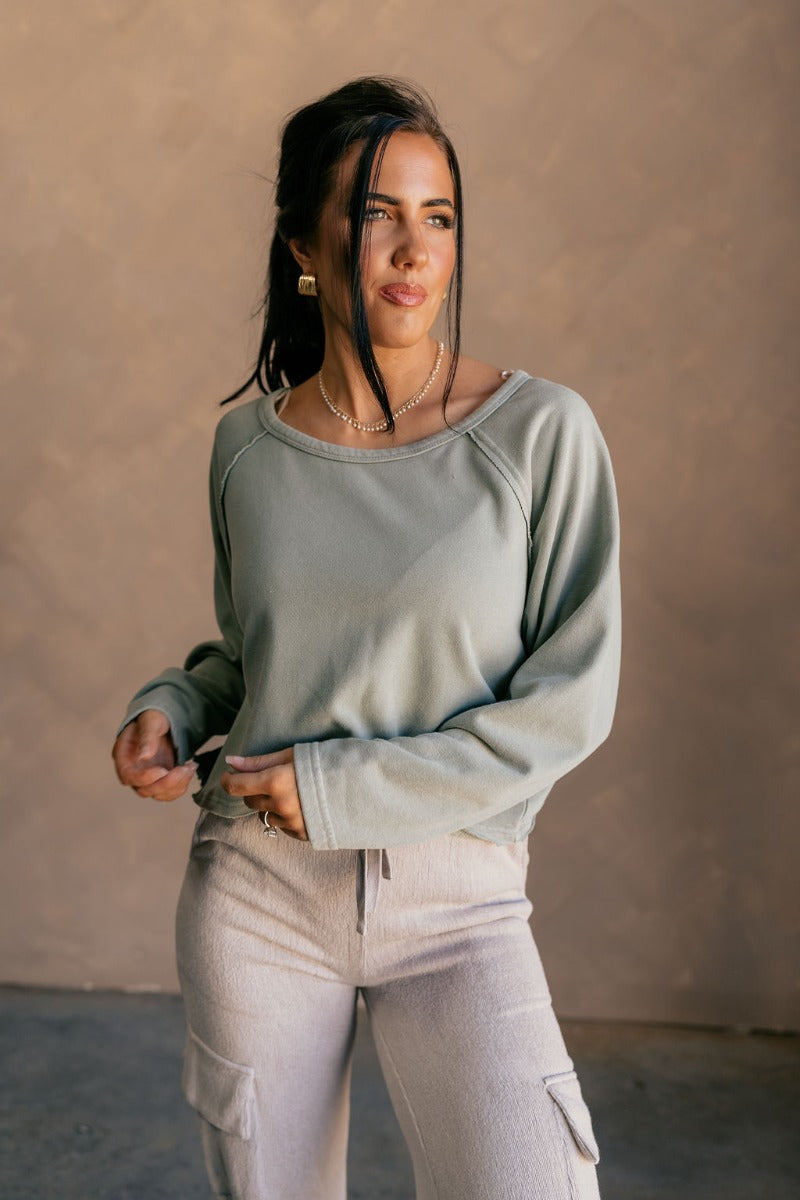 front view of model wearing the Charlotte Sage Green Raglan Sweatshirt that has dusty light sage knit fabric, raw hem details, a round neckline, and long wide sleeves.