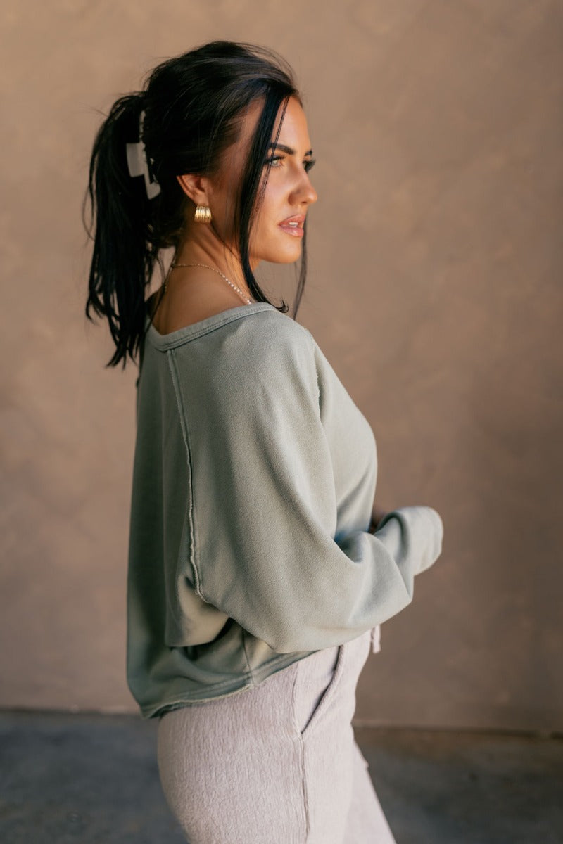 side view of model wearing the Charlotte Sage Green Raglan Sweatshirt that has dusty light sage knit fabric, raw hem details, a round neckline, and long wide sleeves.