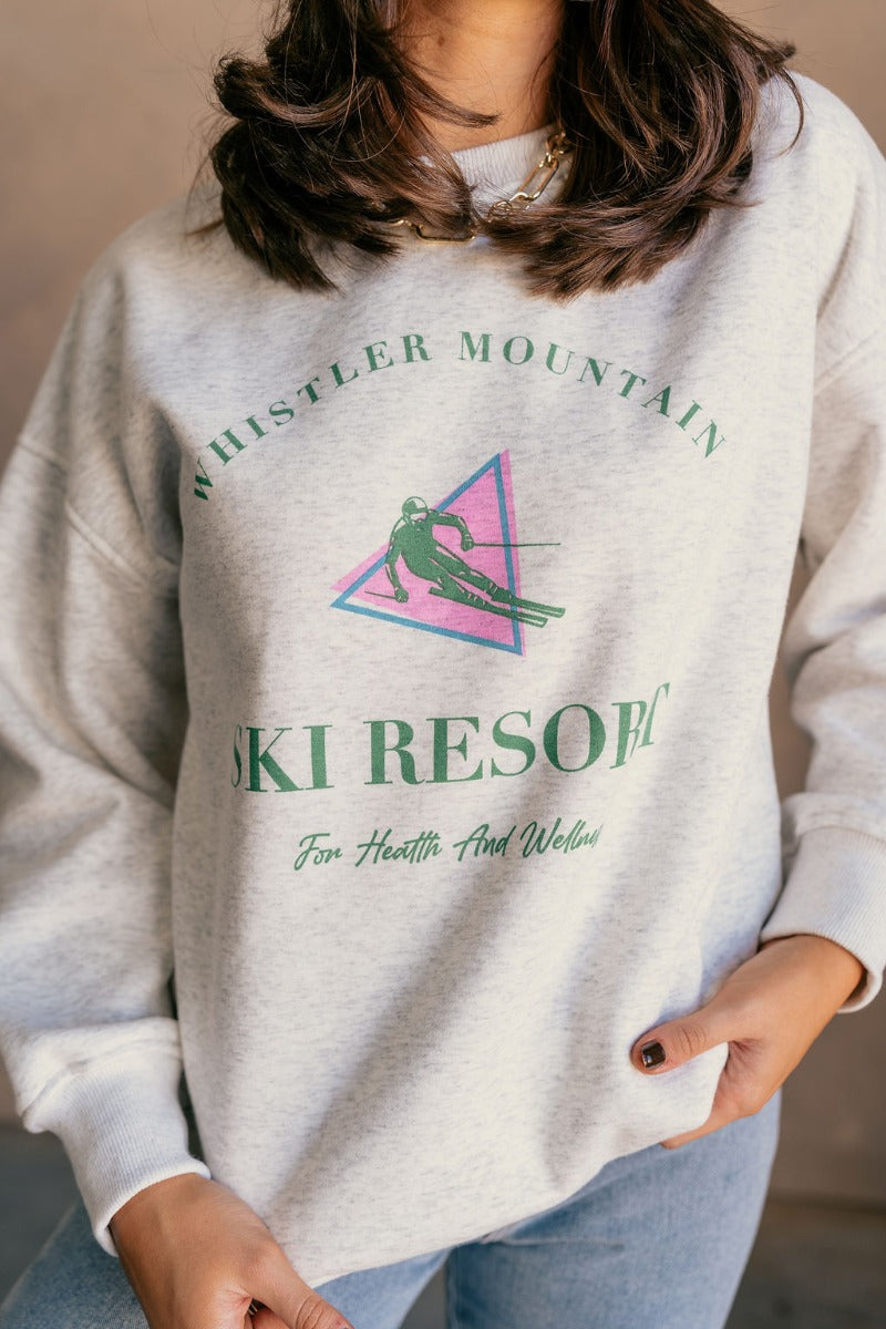 Close up view of model wearing the Whistler Mountain Ski Resort Heather Grey Sweatshirt which features heather grey knit fabric, round neckline, ribbed hem and long sleeves with cuffs. Graphic says "Whistler Mountain Ski Resort" in green "For Health and W