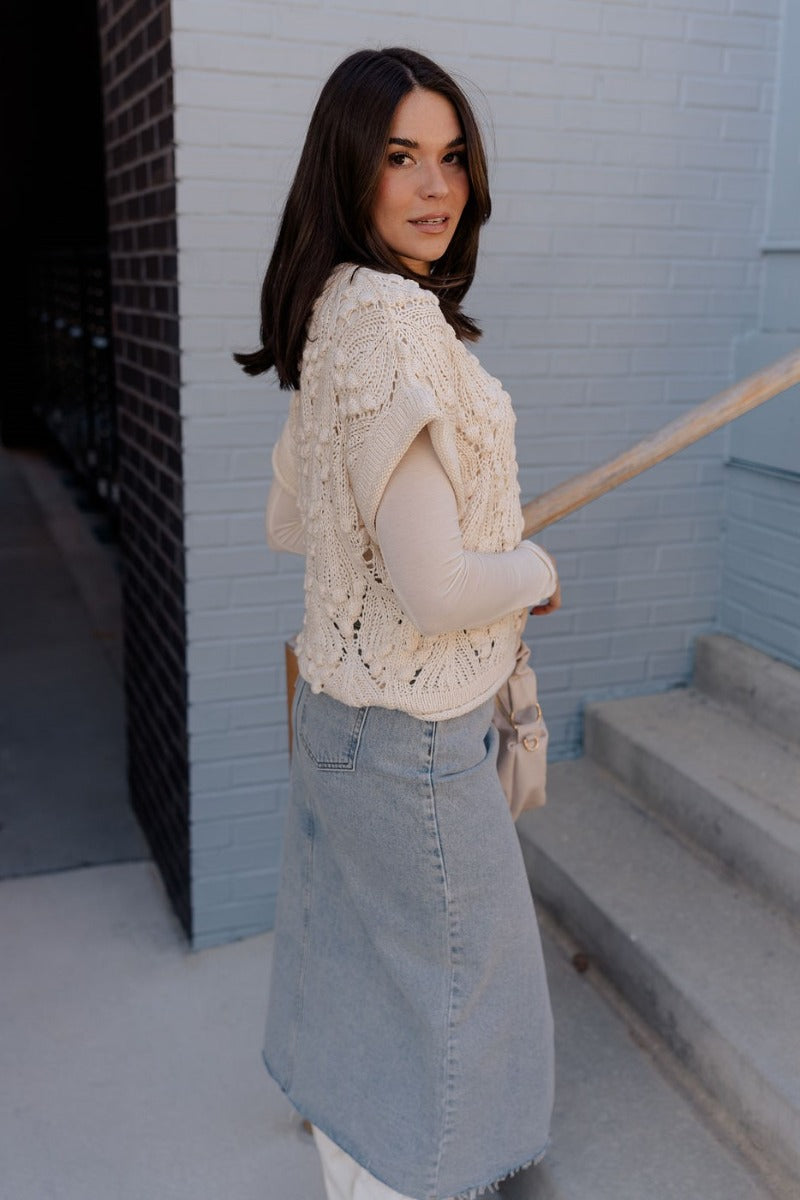 Side view of model wearing the Megan Cream Crochet Short Sleeve Sweater that has cream crochet knit fabric, 3d polka dots, ribbed hem, round neck, and short sleeves. Worn over long sleeve top.