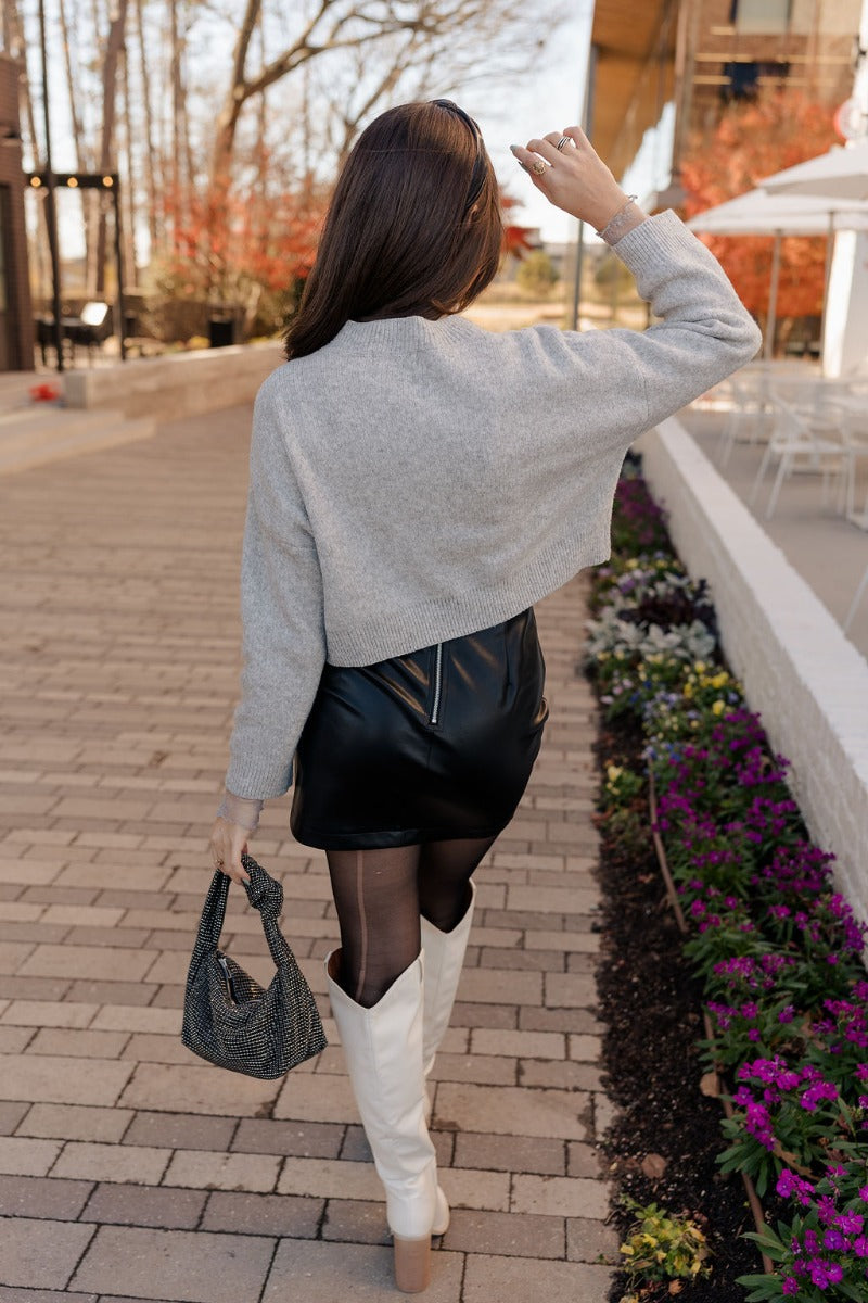 Full body back view of model wearing the Emily Grey Cropped Long Sleeve Sweater that has heather grey knit fabric, ribbed hem, cropped waist, a high neck, dropped shoulders, and long sleeves.