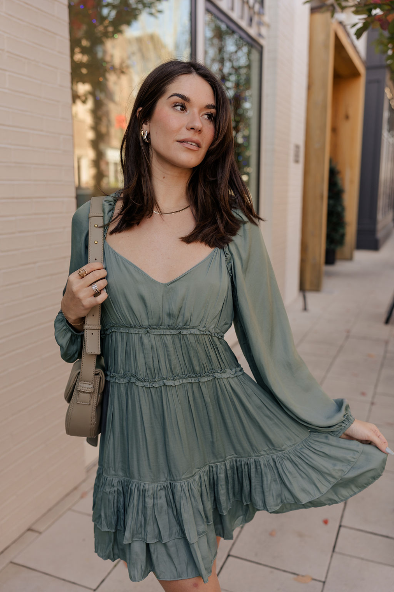 Front view of model wearing the Maya Olive Satin Ruffle Long Sleeve Dress which features olive grey satin fabric, mini length, ruffle details, a three-tier design, olive grey lining, a plunge neckline, long sleeves with elastic cuffs, and a back key hole 