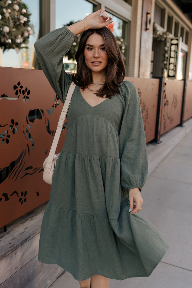 Front view of model wearing the Selene Olive Gauze Midi Long Sleeve Dress features dusty olive gauze fabric, midi length, three tiered design, plunge neckline, smocked back and long sleeves with elastic cuffs.