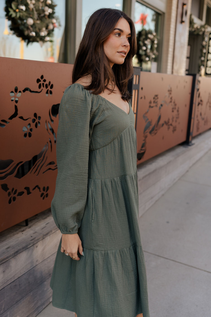 Side view of model wearing the Selene Olive Gauze Midi Long Sleeve Dress features dusty olive gauze fabric, midi length, three tiered design, plunge neckline, smocked back and long sleeves with elastic cuffs.