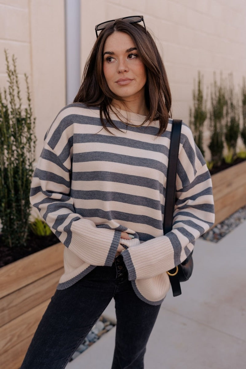 front view of model wearing the Kelly Cream & Grey Striped Long Sleeve Sweater that has cream and grey knit fabric with a striped pattern, a ribbed hem, a round neckline, and long sleeves with ribbed cuffs.