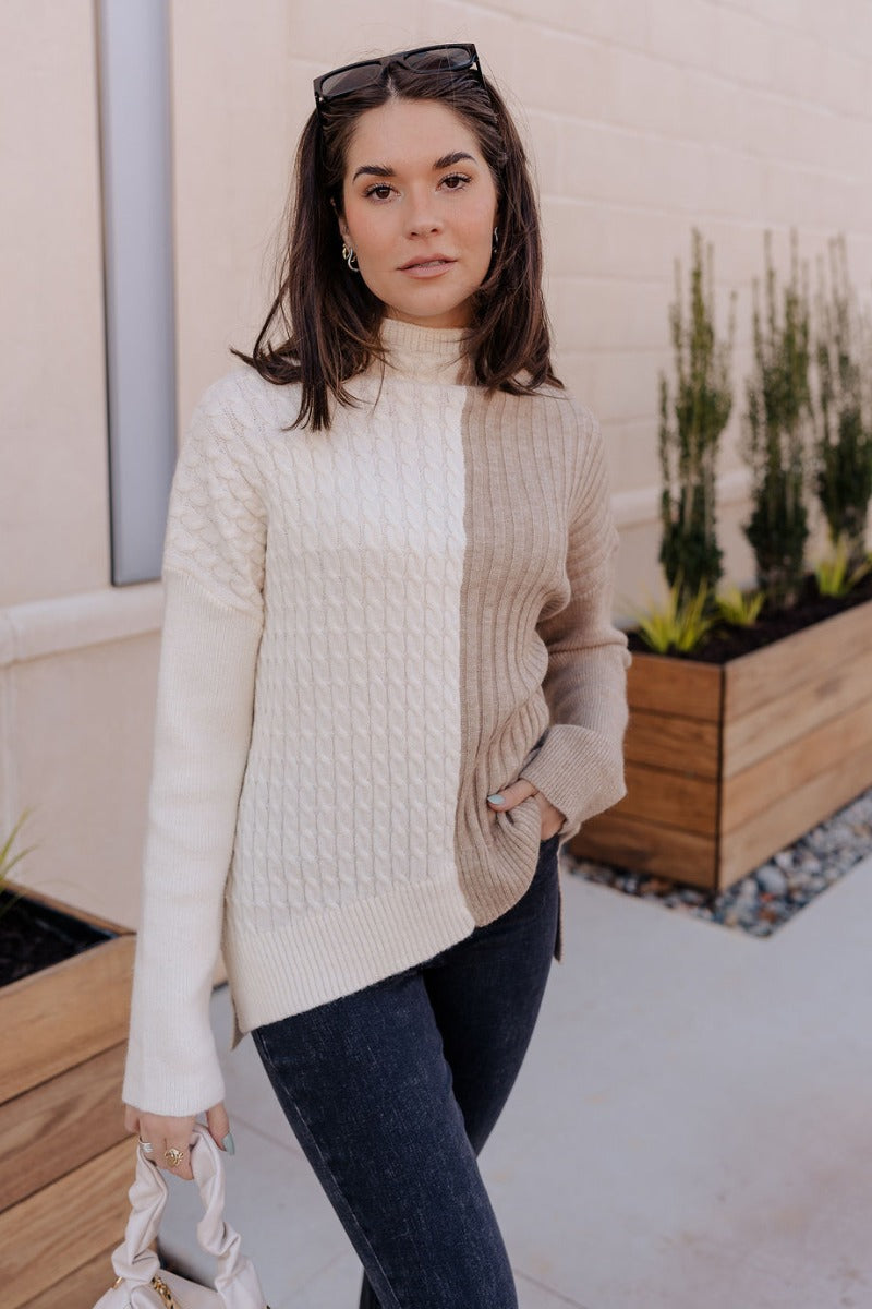 Front view of model wearing the Lillian Taupe & Cream Long Sleeve Sweater which features cream cable knit fabric, taupe ribbed knit fabric, a color block pattern, a ribbed hem, slight slits on each side, a high neckline, and long sleeves with cuffs.