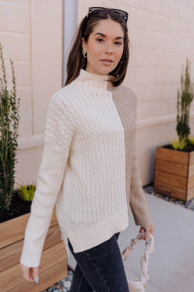 Side view of model wearing the Lillian Taupe & Cream Long Sleeve Sweater which features cream cable knit fabric, taupe ribbed knit fabric, a color block pattern, a ribbed hem, slight slits on each side, a high neckline, and long sleeves with cuffs.