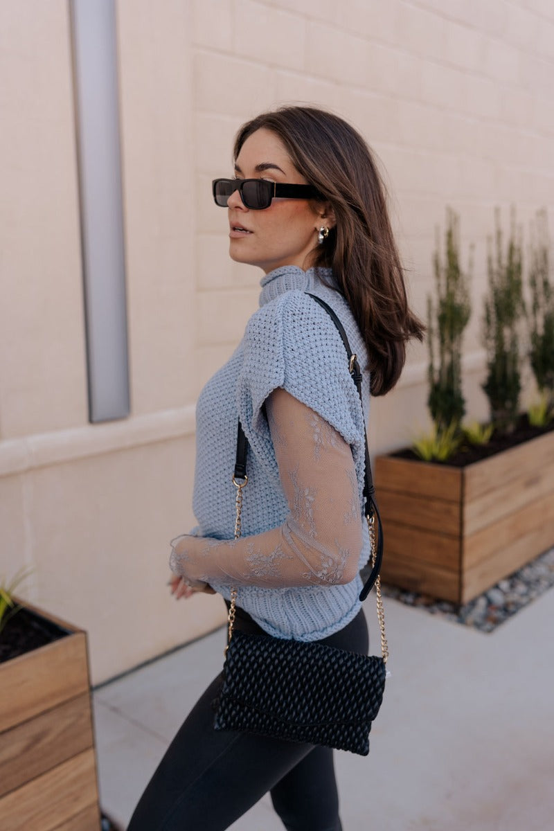 Side view of model wearing the Ryann Baby Blue Sleeveless Sweater that has light blue cable knit fabric, ribbed hem, a high neckline and a sleeveless design.