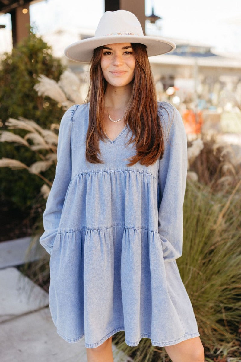 Front view of model wearing the Victoria Denim Babydoll Dress that has light washed denim fabric, a mini-length hem, tiered detailing, a v-neckline, and long balloon sleeves with elastic wrists.