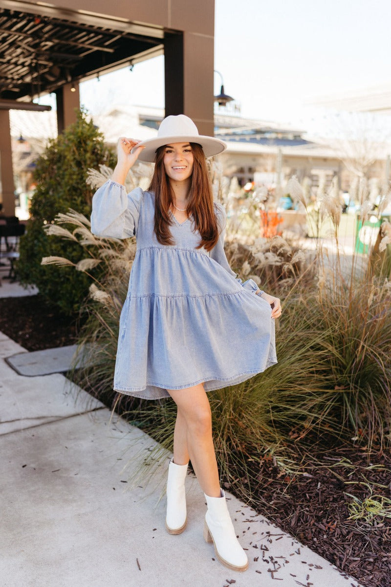 Full body front view of model wearing the Victoria Denim Babydoll Dress that has light washed denim fabric, a mini-length hem, tiered detailing, a v-neckline, and long balloon sleeves with elastic wrists.
