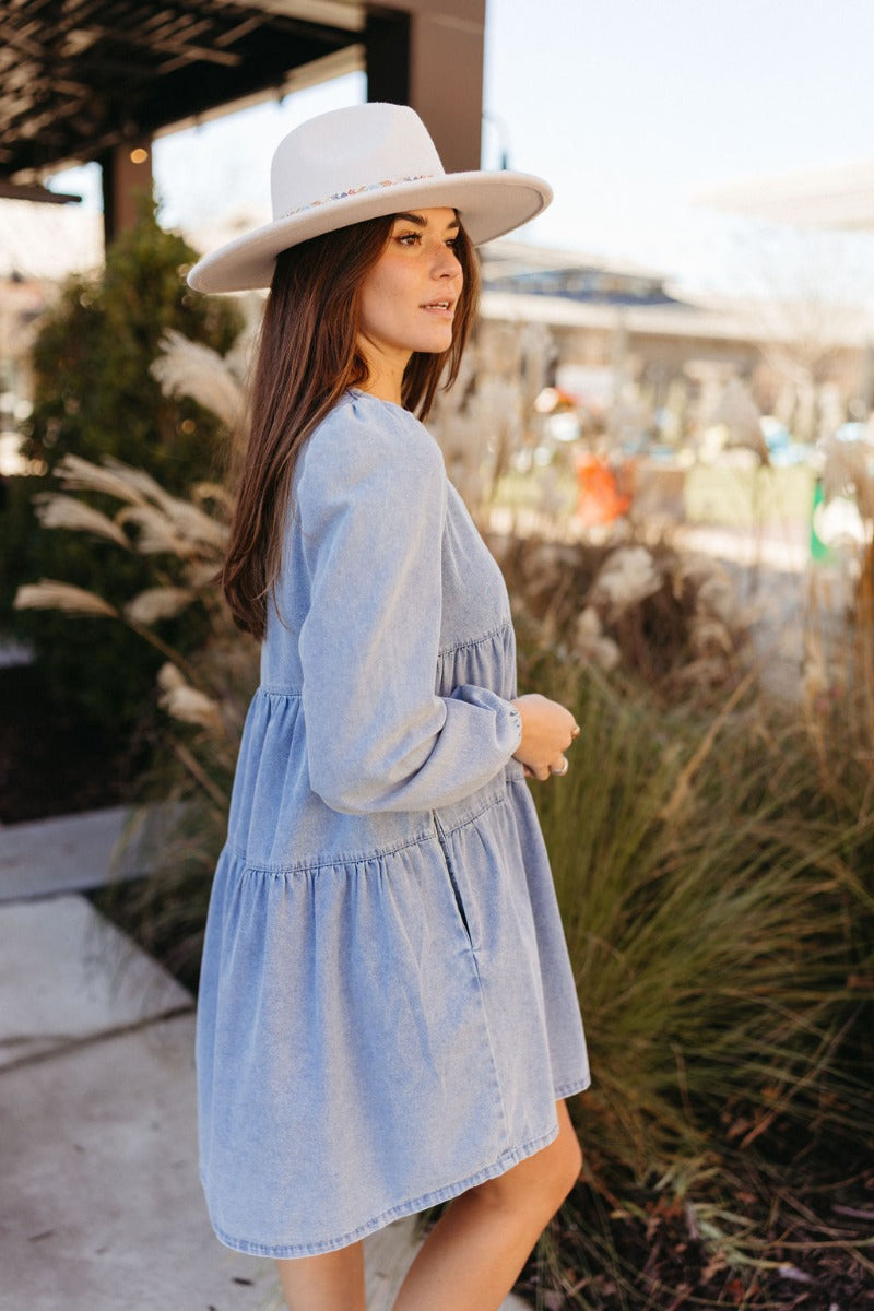 Close side view of model wearing the Victoria Denim Babydoll Dress that has light washed denim fabric, a mini-length hem, tiered detailing, a v-neckline, and long balloon sleeves with elastic wrists.