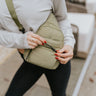 Close up view of the Take A Walk Sling Bag which features olive coloring fabric, two front pockets, large pocket with black zipper and olive adjustable strap for a cross-body look.