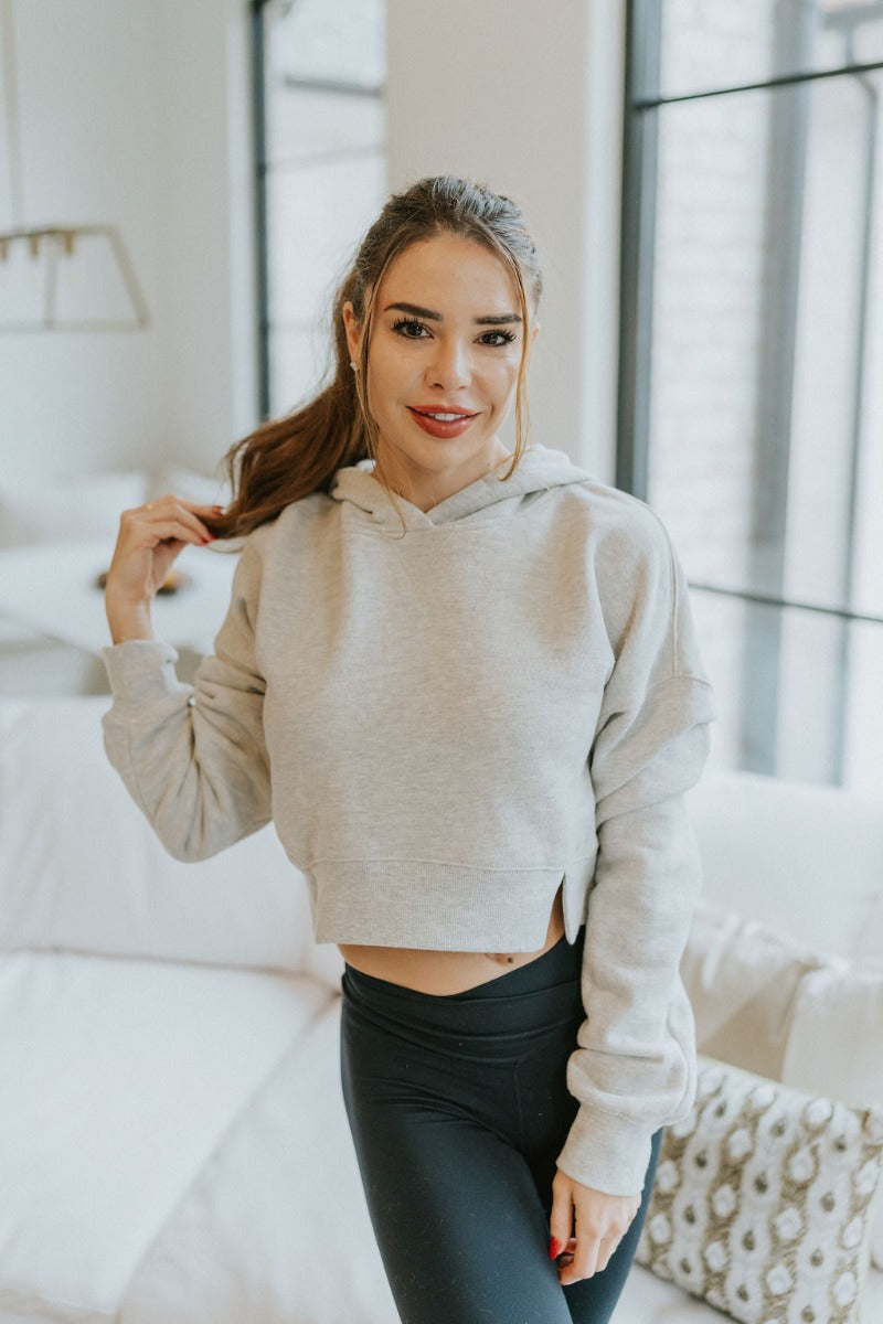 Front view of model wearing the Next Level Hoodie that has heather grey knit fabric, a cropped waist with slits on each side, ribbed trim, a round neckline with a hood, dropped shoulders, and long sleeves