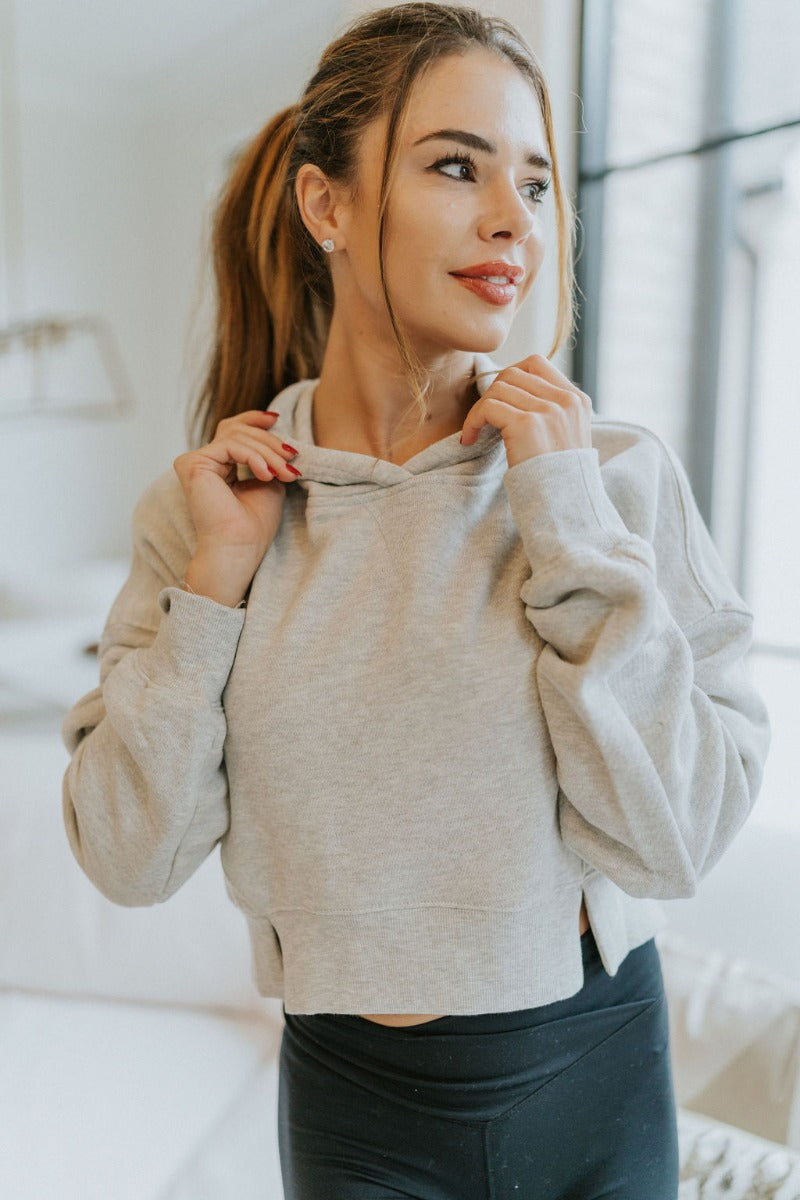 Close front view of model wearing the Next Level Hoodie that has heather grey knit fabric, a cropped waist with slits on each side, ribbed trim, a round neckline with a hood, dropped shoulders, and long sleeves