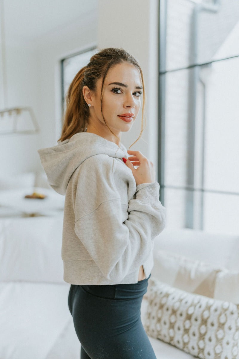 Side view of model wearing the Next Level Hoodie that has heather grey knit fabric, a cropped waist with slits on each side, ribbed trim, a round neckline with a hood, dropped shoulders, and long sleeves