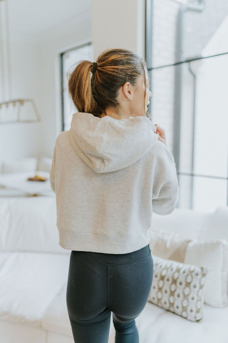 Back view of model wearing the Next Level Hoodie that has heather grey knit fabric, a cropped waist with slits on each side, ribbed trim, a round neckline with a hood, dropped shoulders, and long sleeves