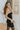 Side view of model wearing the Glam It Up Dress which features black sheen fabric with gold speckles, one shoulder with adjustable spaghetti strap, lining and ruched detail along the entire dress.