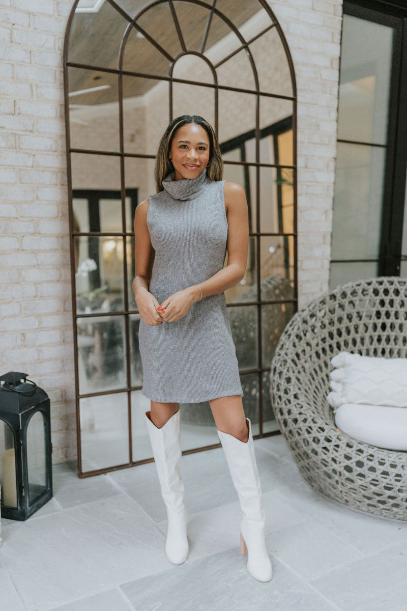Full body front view of model wearing the Call Me Later Sweater Dress that has grey knit fabric, a mini length hem, a loose turtleneck, and a sleeveless body.