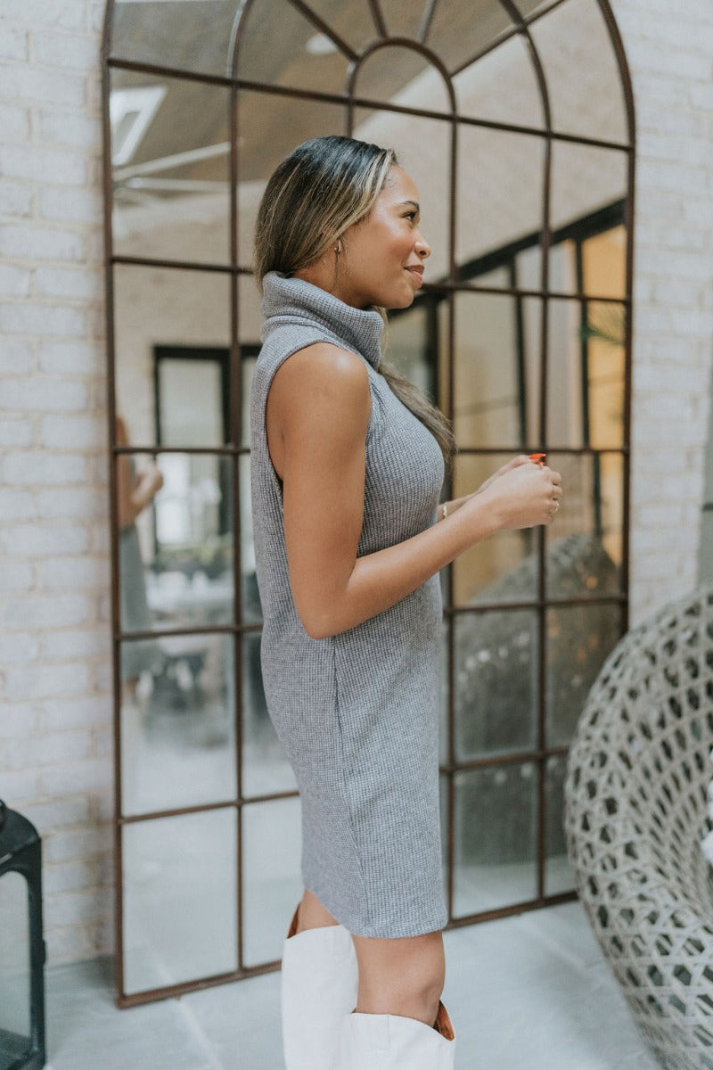 Side view of model wearing the Call Me Later Sweater Dress that has grey knit fabric, a mini length hem, a loose turtleneck, and a sleeveless body.