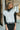 Front view of model wearing the Walk The Line Sweater Vest in White that has white knit fabric, ribbed trim with black stripes, a v-neckline and a sleeveless body