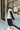 Side view of model wearing the Walk The Line Sweater Vest in White that has white knit fabric, ribbed trim with black stripes, a v-neckline and a sleeveless body