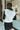 Back view of model wearing the Walk The Line Sweater Vest in White that has white knit fabric, ribbed trim with black stripes, a v-neckline and a sleeveless body