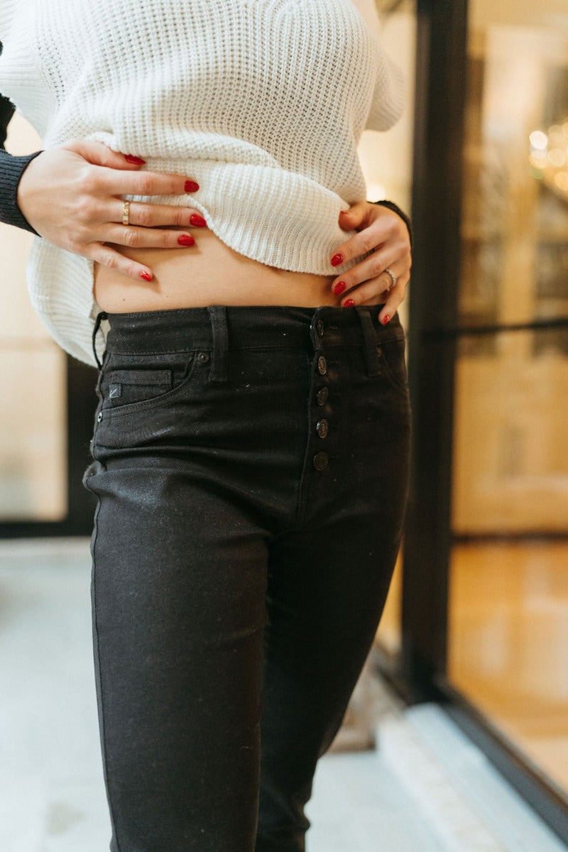 Close front view of model wearing the KanCan: Trend Report Jeans that have black sheen denim fabric, a 5-button closure, two pockets, back pockets, belt loops, high-rise waist, and cropped flare legs with a raw hem