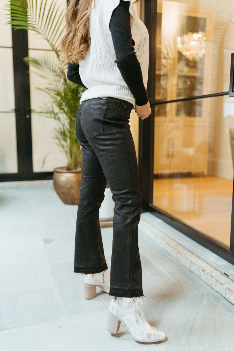 Side view of model wearing the KanCan: Trend Report Jeans that have black sheen denim fabric, a 5-button closure, two pockets, back pockets, belt loops, high-rise waist, and cropped flare legs with a raw hem