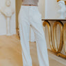 Front view of model wearing the East Side Pants that have cream fabric, front pockets, side leg pockets, a zipper and button, belt loops, an elastic back waist, back pockets, and wide legs.