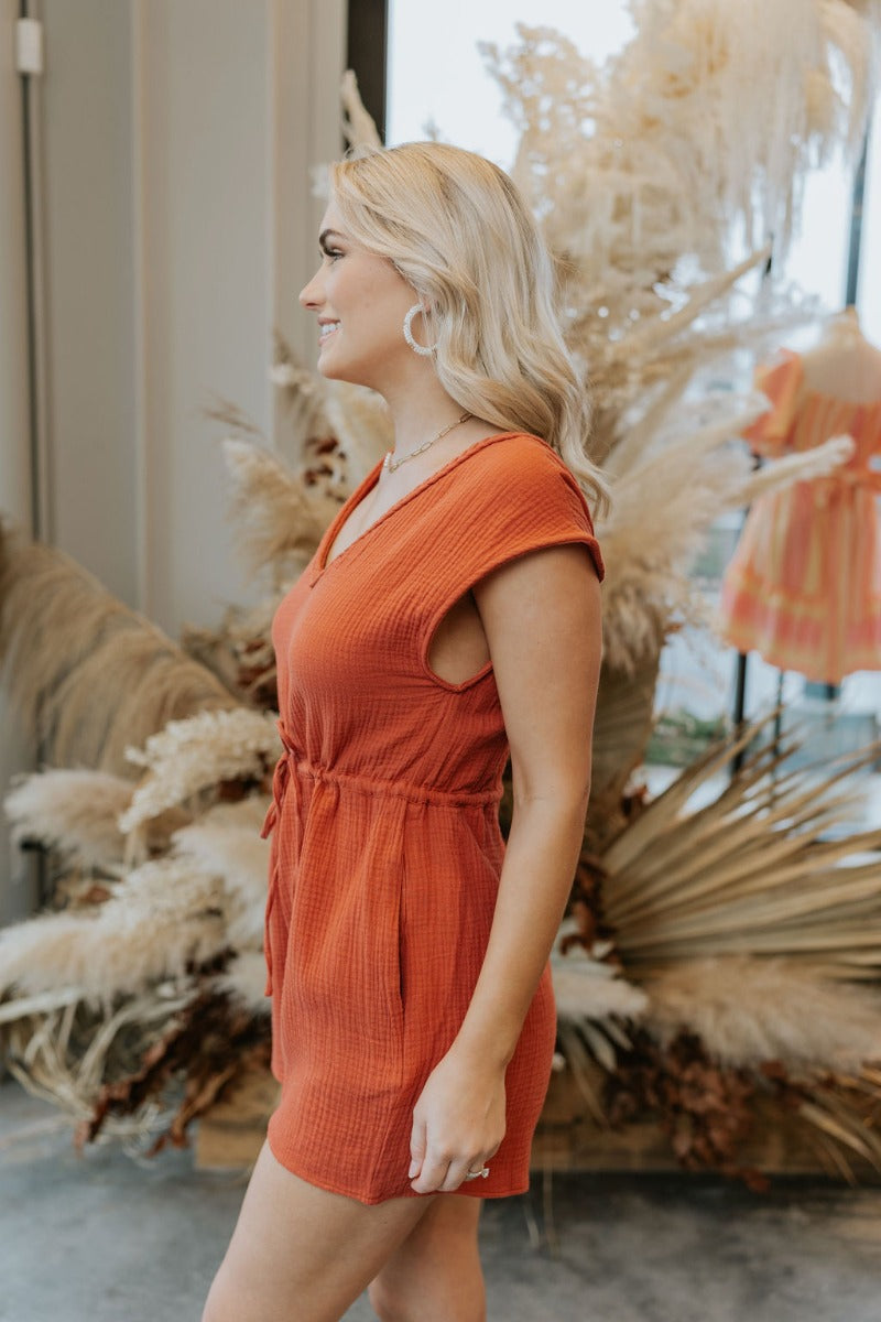 Side view of model wearing the Sahara Views Romper which features rust gauze fabric, pockets on the side, a drawstring around the waistline, a v-neckline with roping details, a slight open back with rope details, and a sleeveless design.