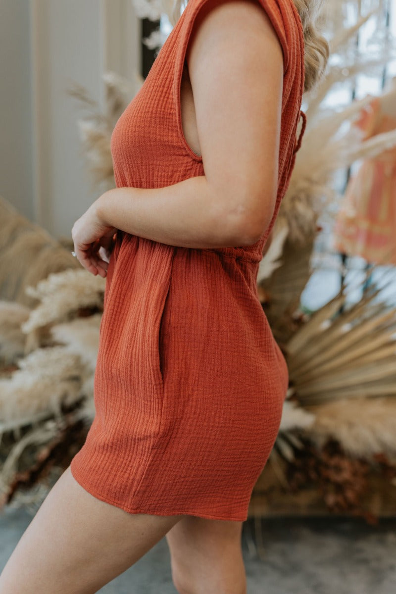 Close up/side view of model wearing the Sahara Views Romper which features rust gauze fabric, pockets on the side, a drawstring around the waistline, a v-neckline with roping details, a slight open back with rope details, and a sleeveless design.