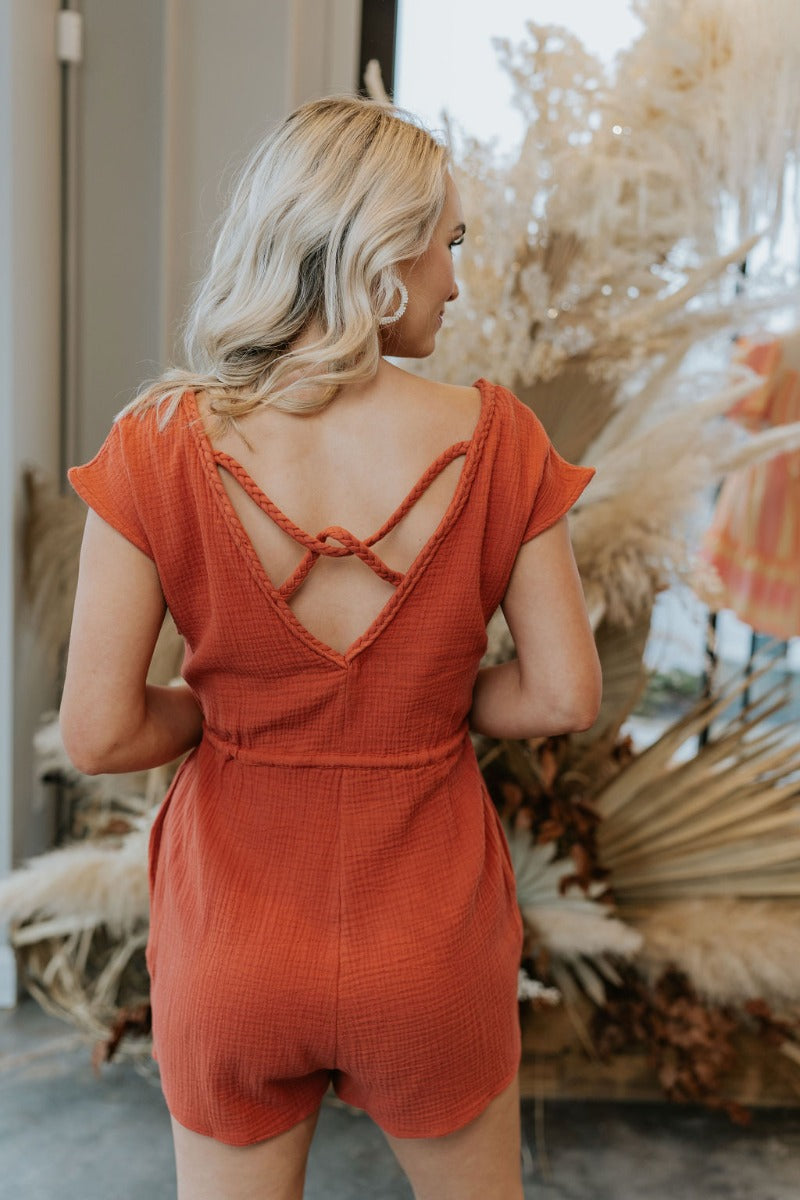 Back view of model wearing the Sahara Views Romper which features rust gauze fabric, pockets on the side, a drawstring around the waistline, a v-neckline with roping details, a slight open back with rope details, and a sleeveless design.