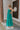 Side view of model wearing the No More Wishing Dress which features kelly green fabric with a lining, a tiered midi-length skirt, a ruched neckline, adjustable thick straps, and a smocked back.