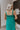 Close up/Back view of model wearing the No More Wishing Dress which features kelly green fabric with a lining, a tiered midi-length skirt, a ruched neckline, adjustable thick straps, and a smocked back.