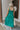 Back view of model wearing the No More Wishing Dress which features kelly green fabric with a lining, a tiered midi-length skirt, a ruched neckline, adjustable thick straps, and a smocked back.