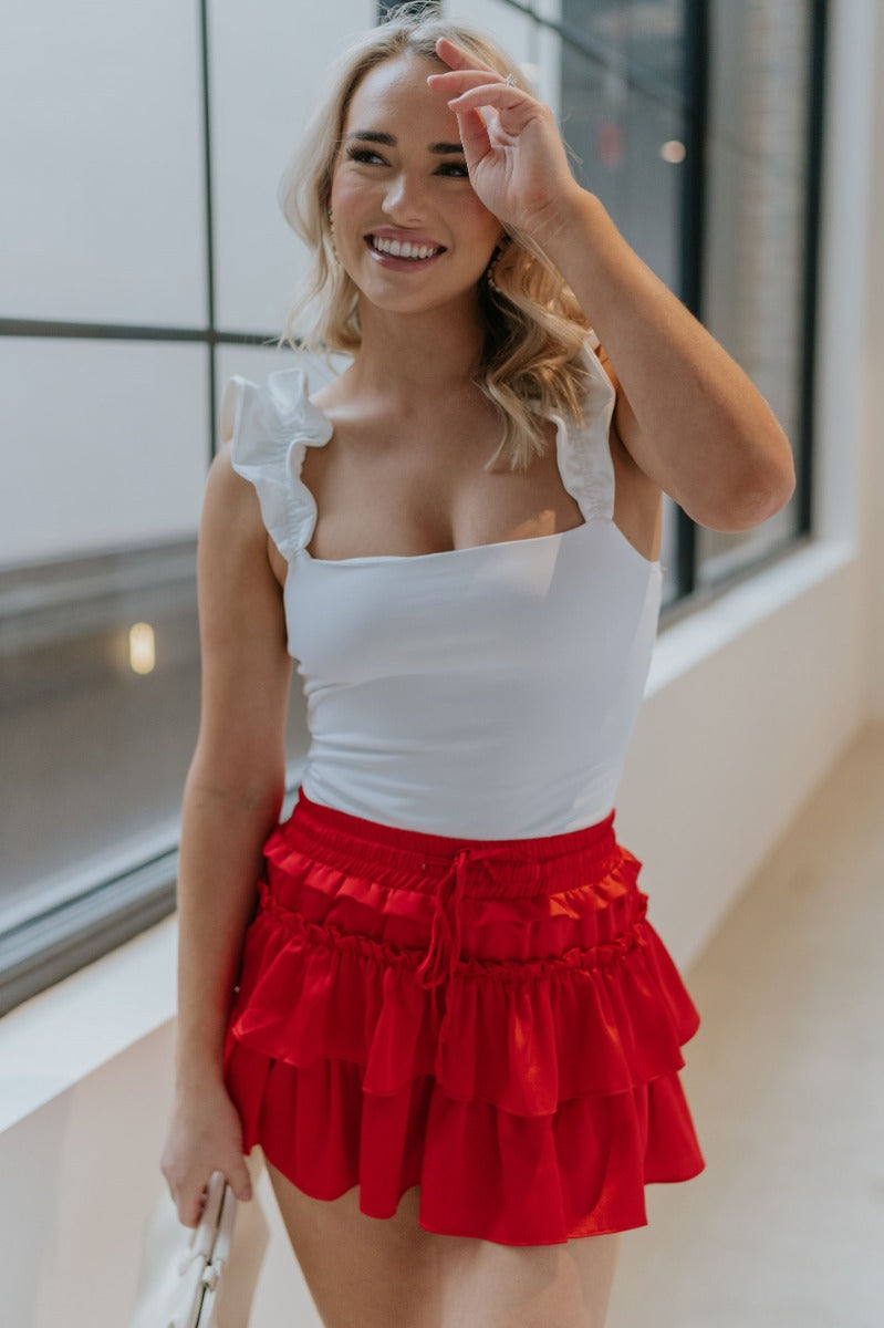 Front view of model wearing the It's Complicated Satin Skort in Red that has red satin fabric, a tiered body with ruffle details, an elastic drawstring waist, and shorts lining.