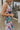 Close side view of model wearing the In Bloom Midi Dress that has white fabric with a purple, blue, green and pink watercolor floral design, ruching, a v-neck, thick straps, a a side slit, and a lining